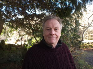 A photograph of a man with short grey hair, wearing a dark jumper, there is a tree in the background