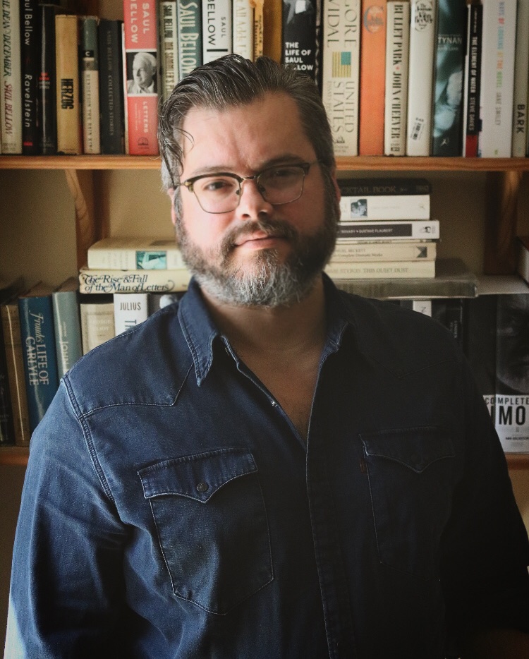 A man with glasses and a beard, with short dark hair, posed in front of a bookcase.