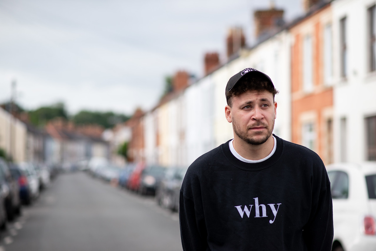 A man with a baseball cap on. He is wearing a sweatshirt with the word 'why'. The background is a blurred terraced street