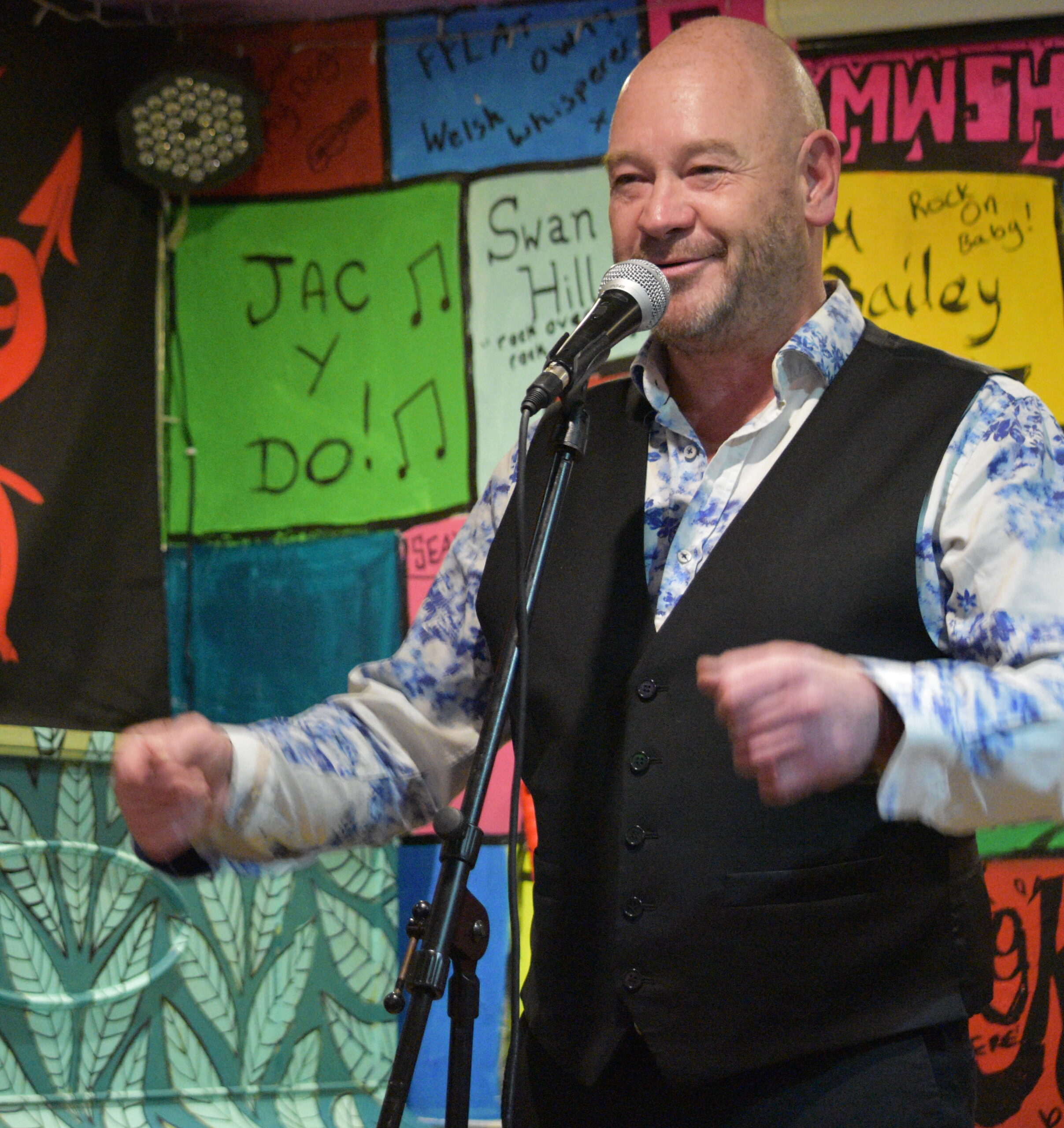 A bald man wearing a brightly patterned long sleeve shirt and dark waistcoat standing in front of a microphone. The background is colourful posters.