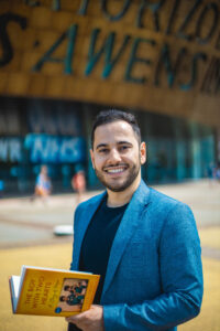 A man with short hair, wearing a bright blue blazer over a dark t-shirt. Holding an open book with a yellow cover. The background is blurred version of the Wales Millennium Centre in Cardiff. 