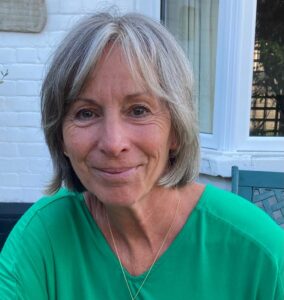 A woman with grey hair in a bob, wearing a green t-shirt. There is a white wall and a window in the background. 