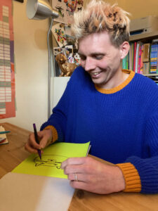 A man with blonde spiked hair, wearing a bright blue and orange jumper, they are drawing a cat face on yellow paper. 
