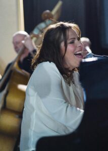 A woman with long brown hair, wearing a white shirt. She is sat at a piano and singing. In the background is a man playing a double bass.