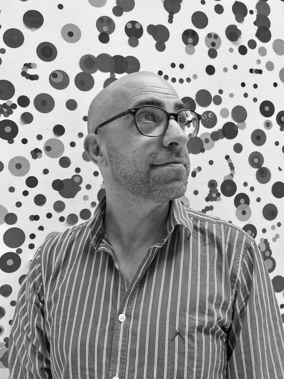 A greyscale photo of a bald man wearing glasses and a stripped shirt. The background is various sized circles