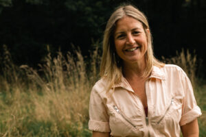The photograph was taken outside. To the right of the image - A woman with shoulder length blonde hair. She is facing the camera and smiling. She is wearing a light colour top with short sleeves and zip detailing. The background is a blurred scene of high grass and trees. 