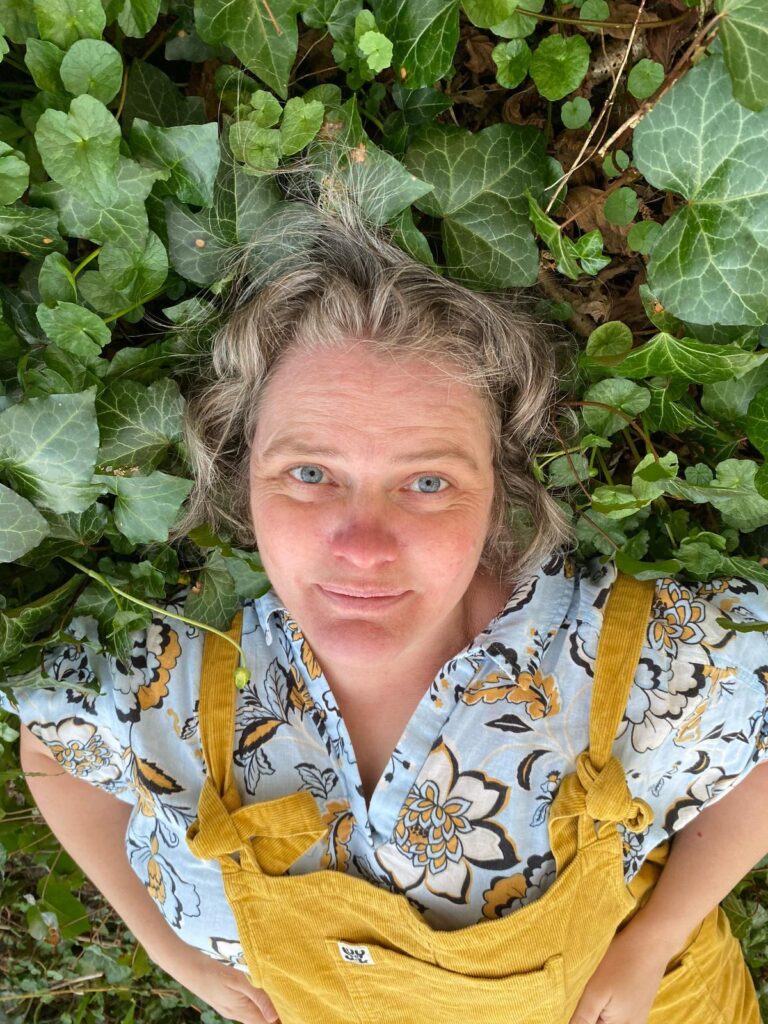 A photograph of a person wearing yellow dungarees over a patterned top. They are lying on a pile of green leaves.