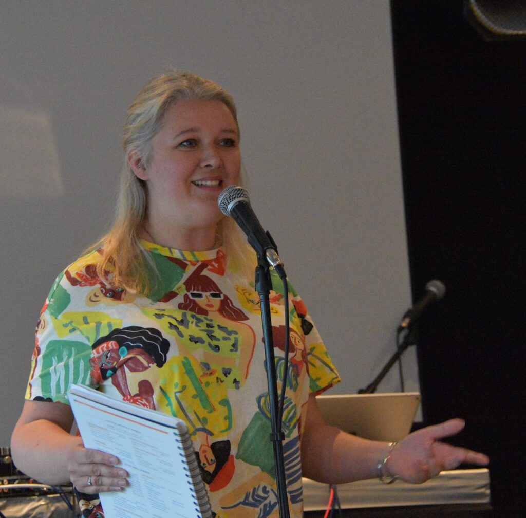 A blonde woman wearing a brightly patterned top, stood in front of a microphone.